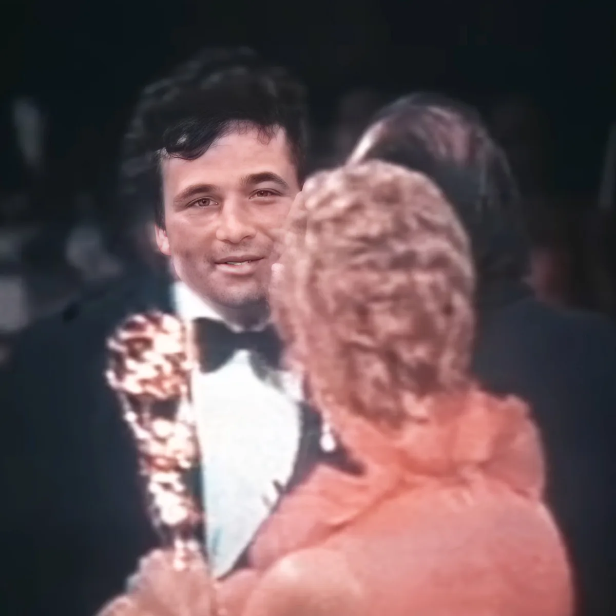 A man in a tuxedo holds an Oscar statue while embracing a woman in a pink dress.