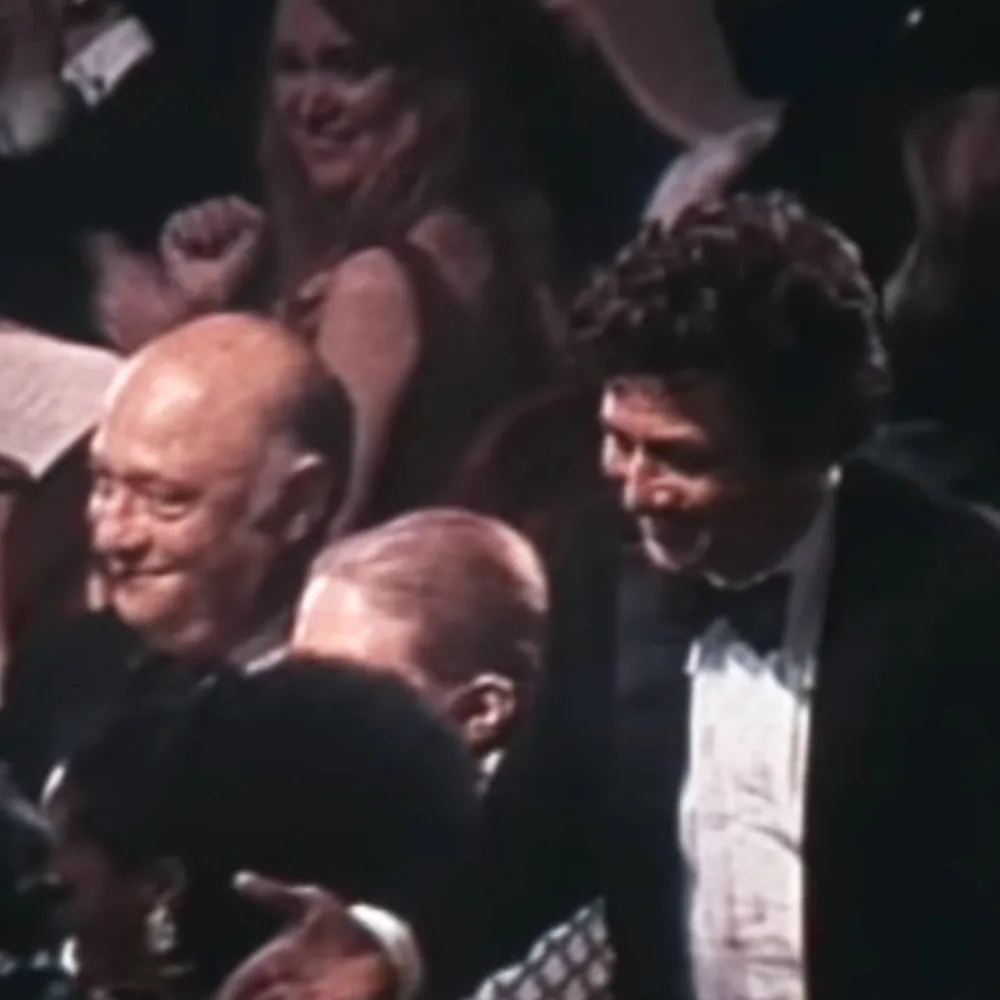 A man in a tuxedo is smiling and clapping, surrounded by other people in formal attire.