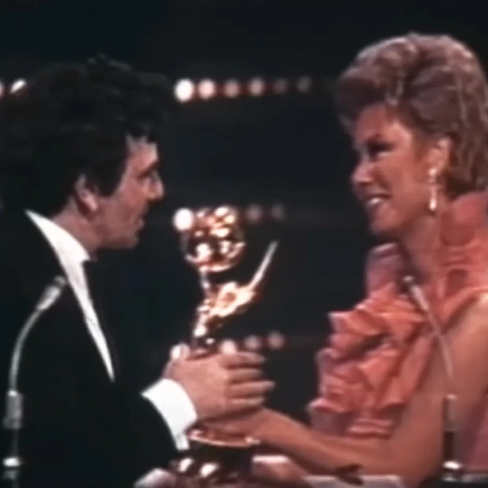 A man in a tuxedo and a woman in a pink dress hold a golden Emmy award.