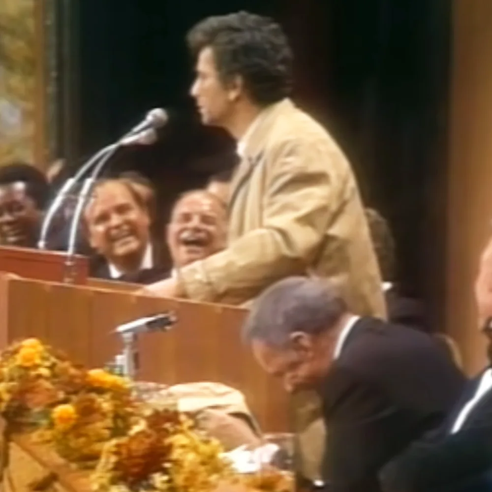 A man in a beige jacket stands at a podium, speaking to an audience, with a woman smiling in the background.
