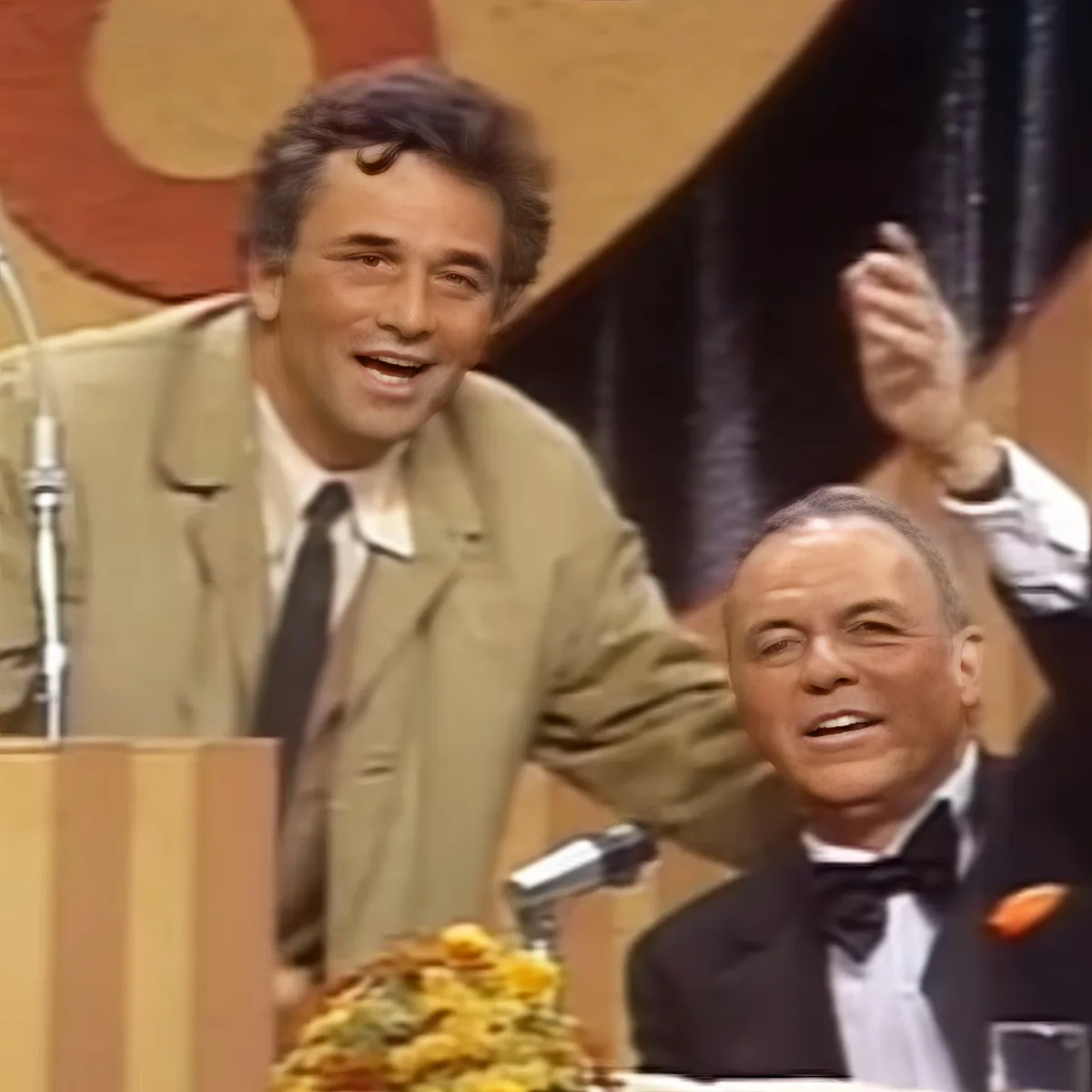 A man in a tan suit stands at a podium, smiling, with another man in a tuxedo seated beside him, raising his hand.