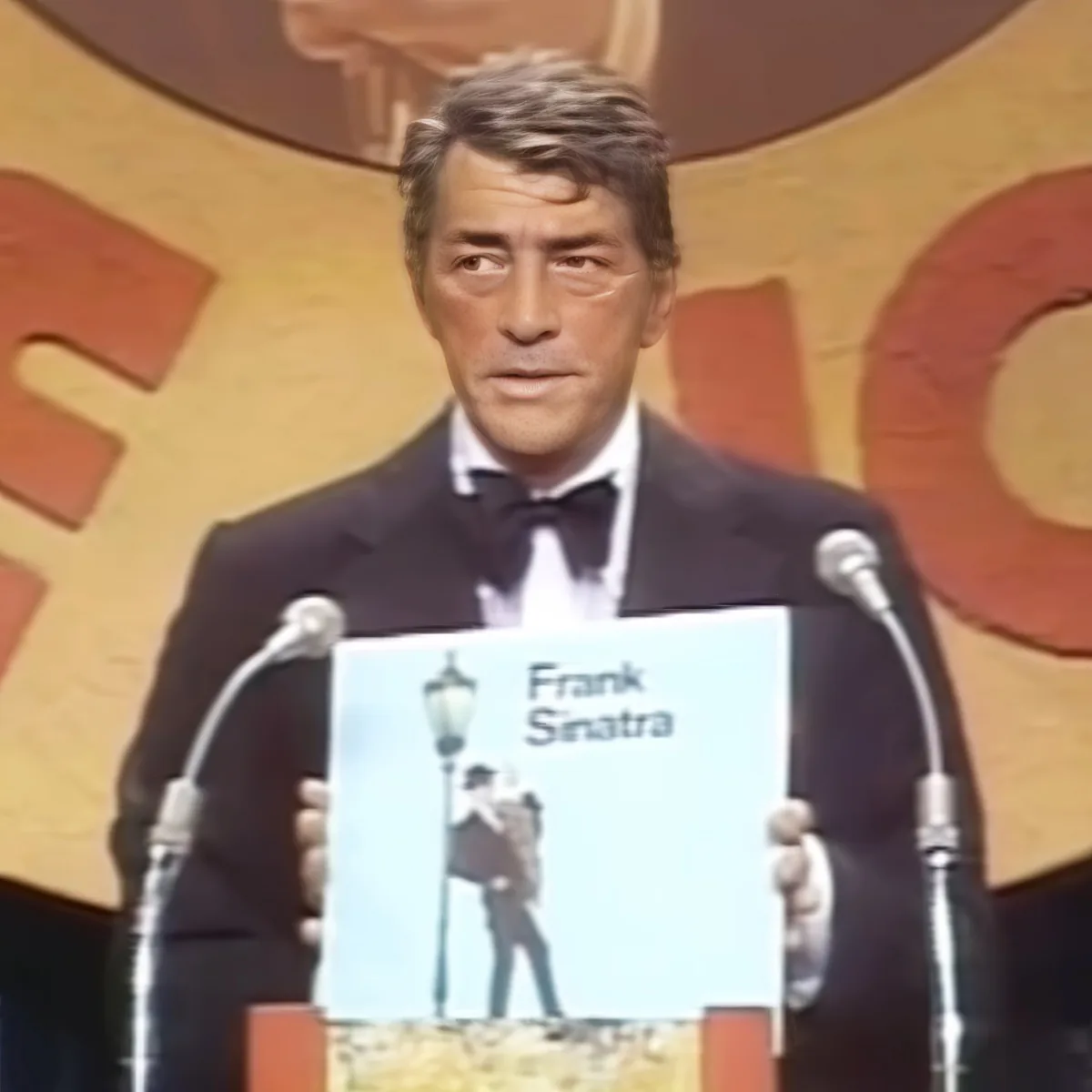 A man in a tuxedo stands at a podium, holding a Frank Sinatra album cover.