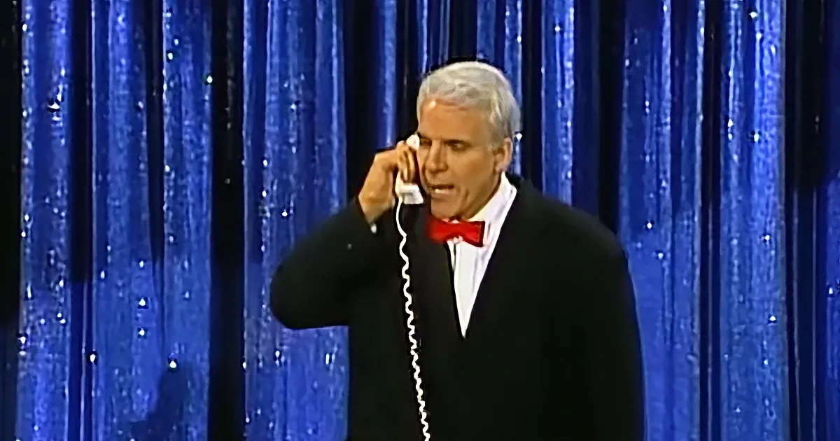 A man in a tuxedo and bow tie is holding a phone to his ear, standing in front of a blue curtain.