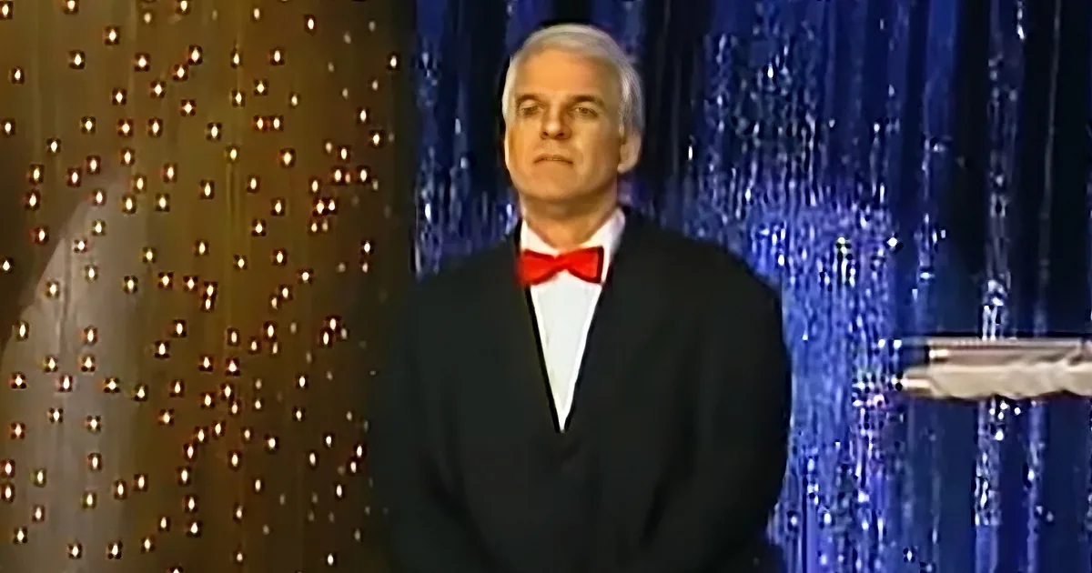 A man in a tuxedo with a red bow tie stands on stage with a glittery backdrop.