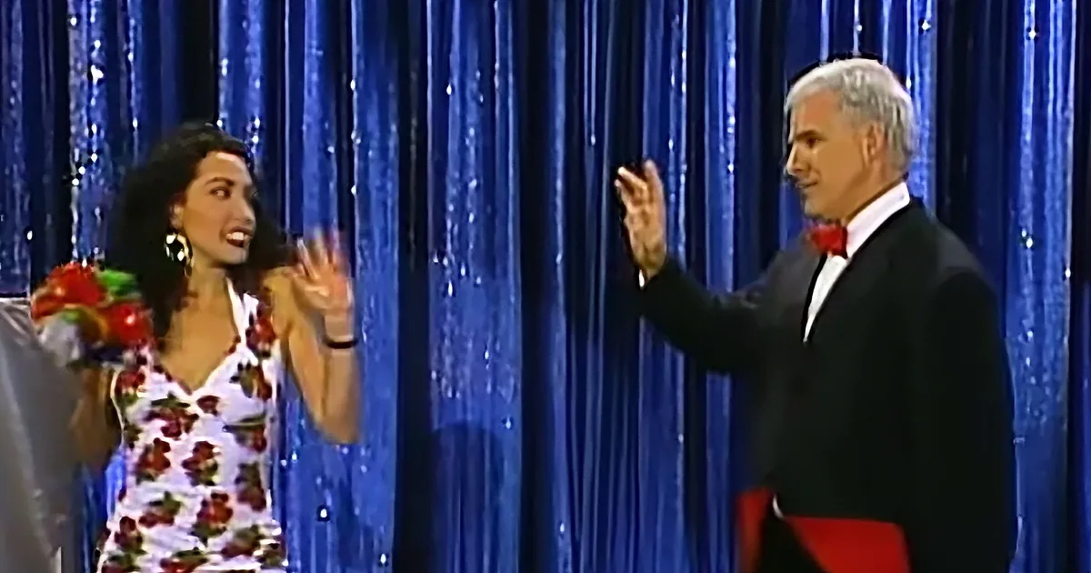 A woman in a floral dress holds flowers, gesturing, while a man in a tuxedo raises his hand, both on stage.