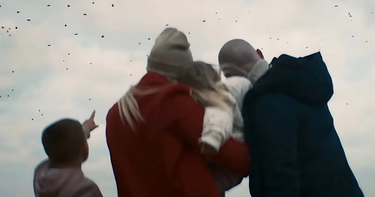 A family of four, including a child, stands together, looking up at birds flying in the sky.