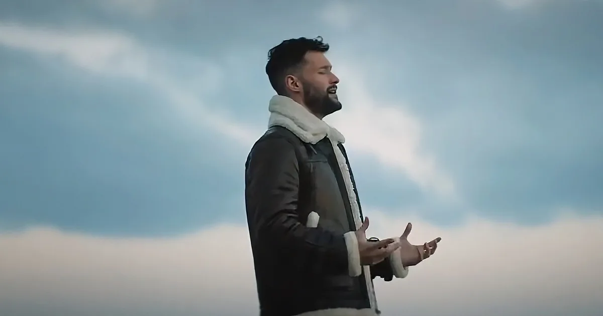 A man in a leather jacket and scarf stands with hands outstretched against a cloudy sky.