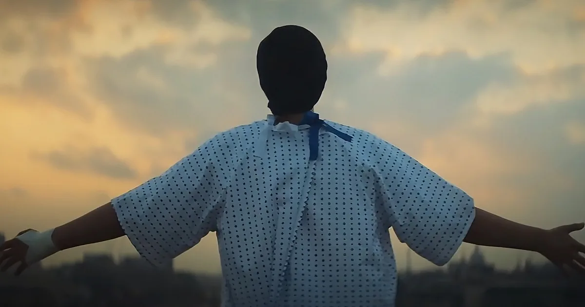 A person in a white polka-dot shirt with a blue bow and a black mask stands with arms outstretched against a cloudy sky.