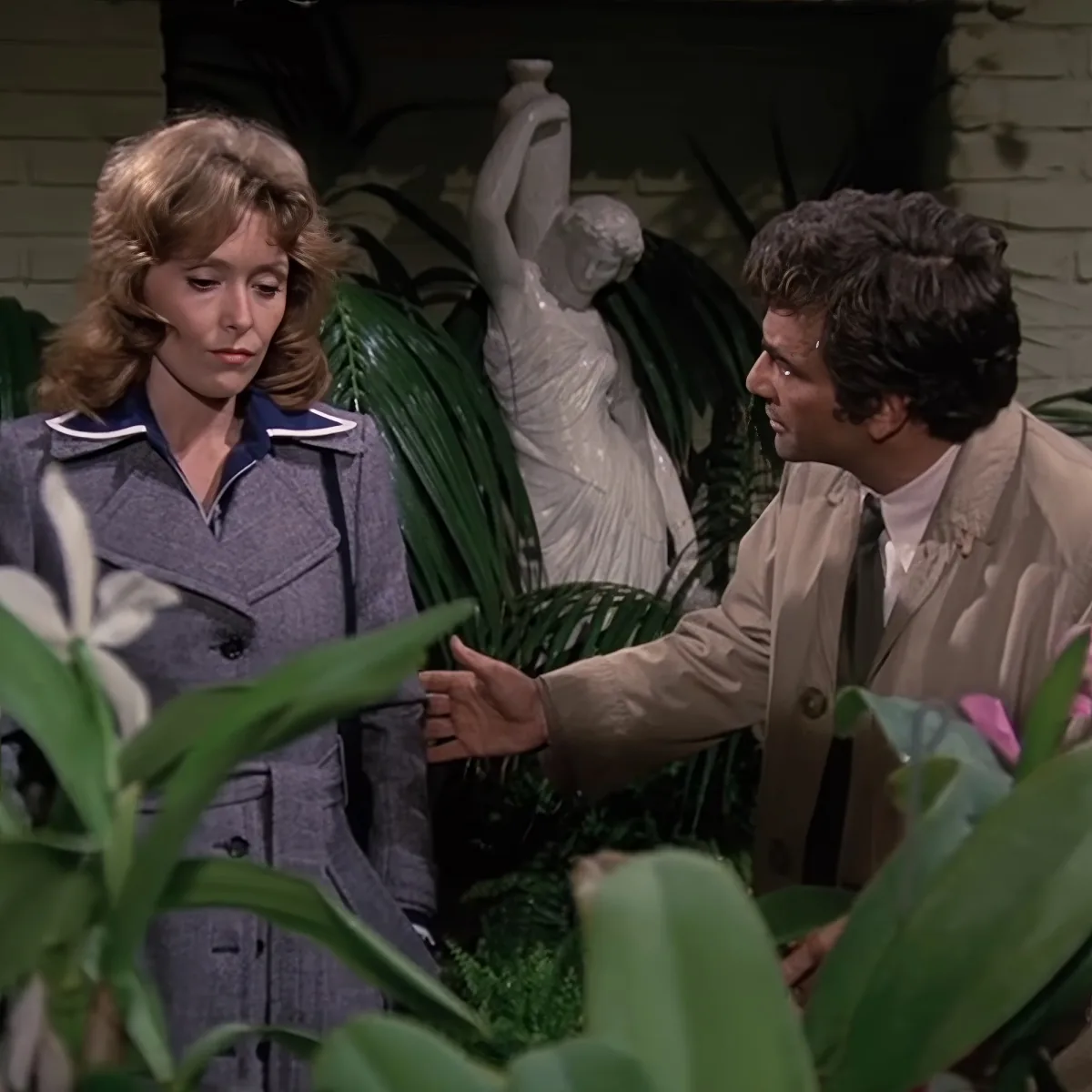 A man in a beige coat gestures towards a woman in a gray jacket amidst greenery and a statue.