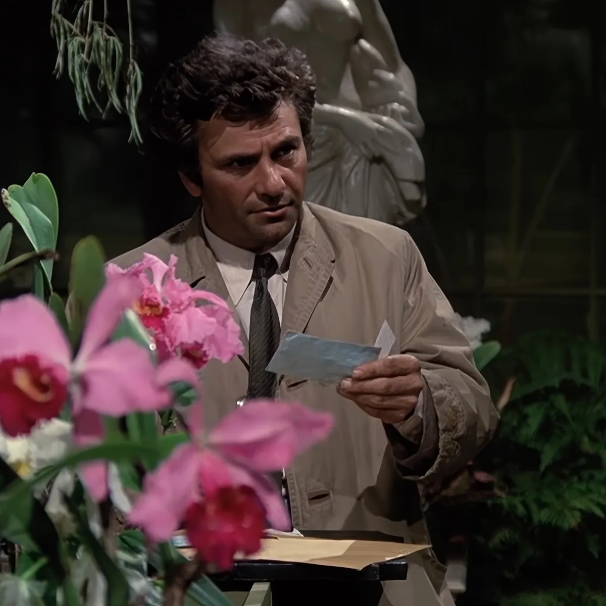 A man in a beige suit holds a piece of paper, surrounded by pink orchids and a statue in the background.