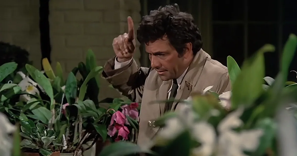 A man in a tan jacket points upwards while surrounded by plants and flowers.