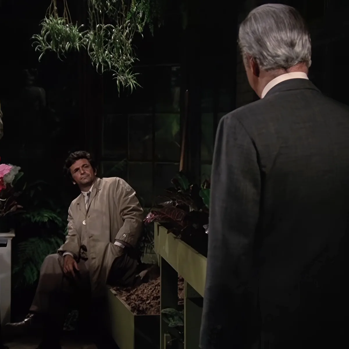 A man in a tan suit leans against a wall in a dimly lit room with plants and another man in a suit facing him.