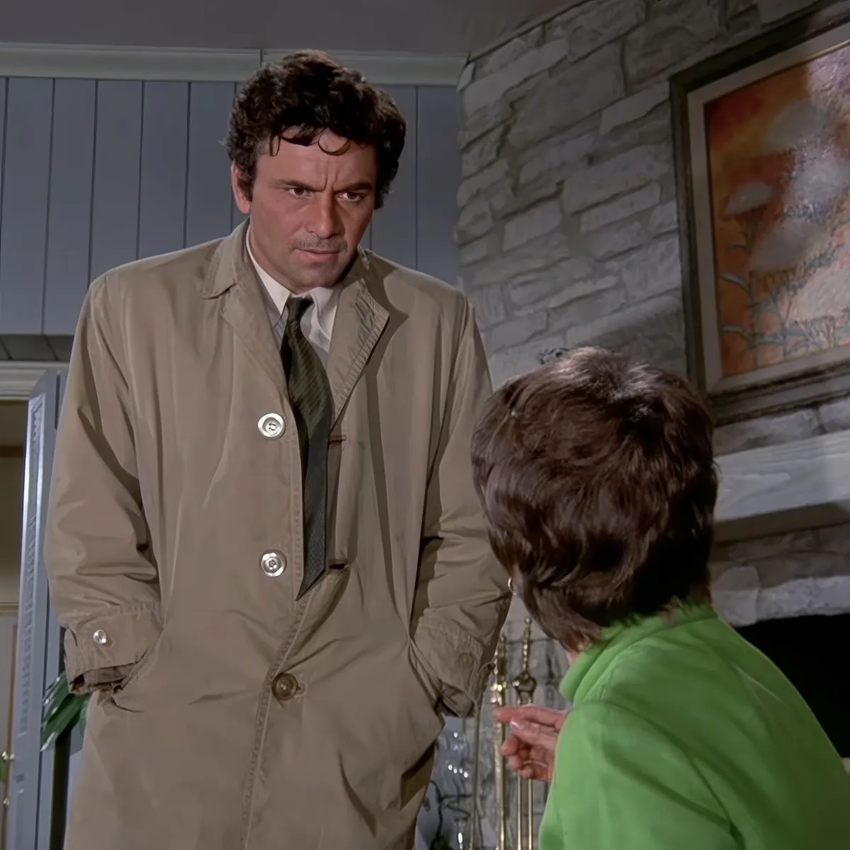 A man in a beige trench coat and tie stands in a room, looking concerned, while a woman in a green top stands opposite him.