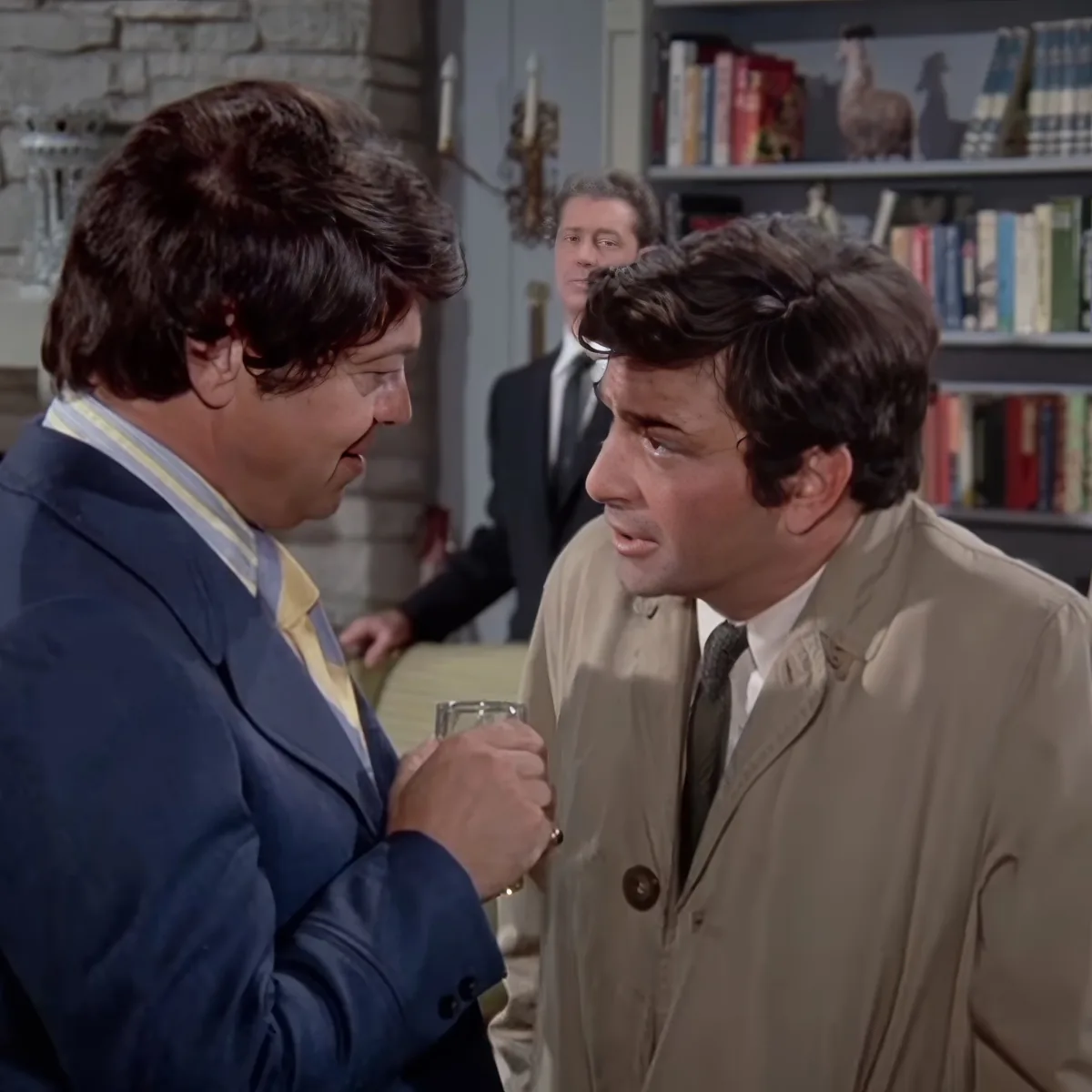 A man in a blue suit holds a glass, interacting with another man in a beige coat in a room with bookshelves.