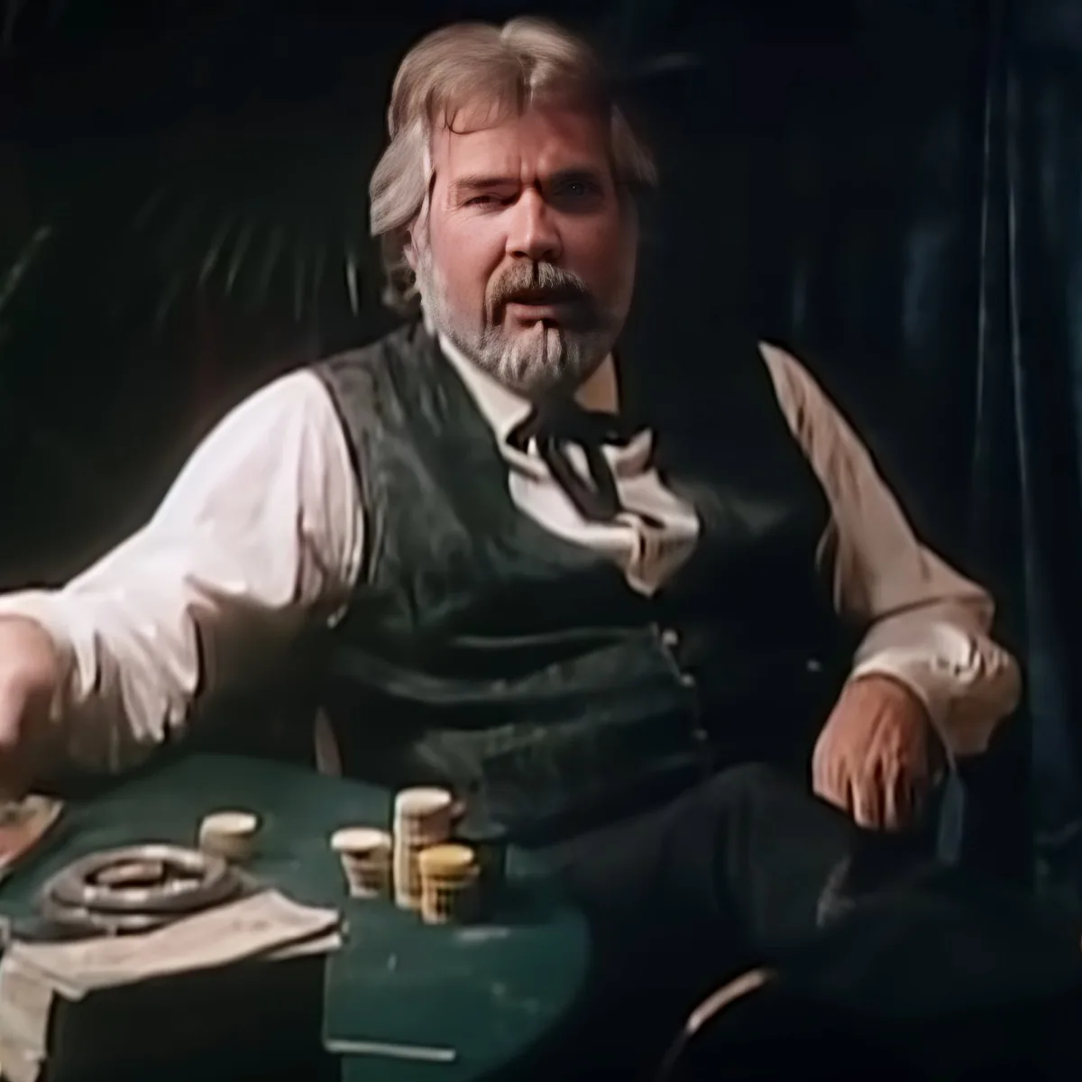 A man with a beard and white hair, wearing a vest and bow tie, sits at a table with coins and a green object.