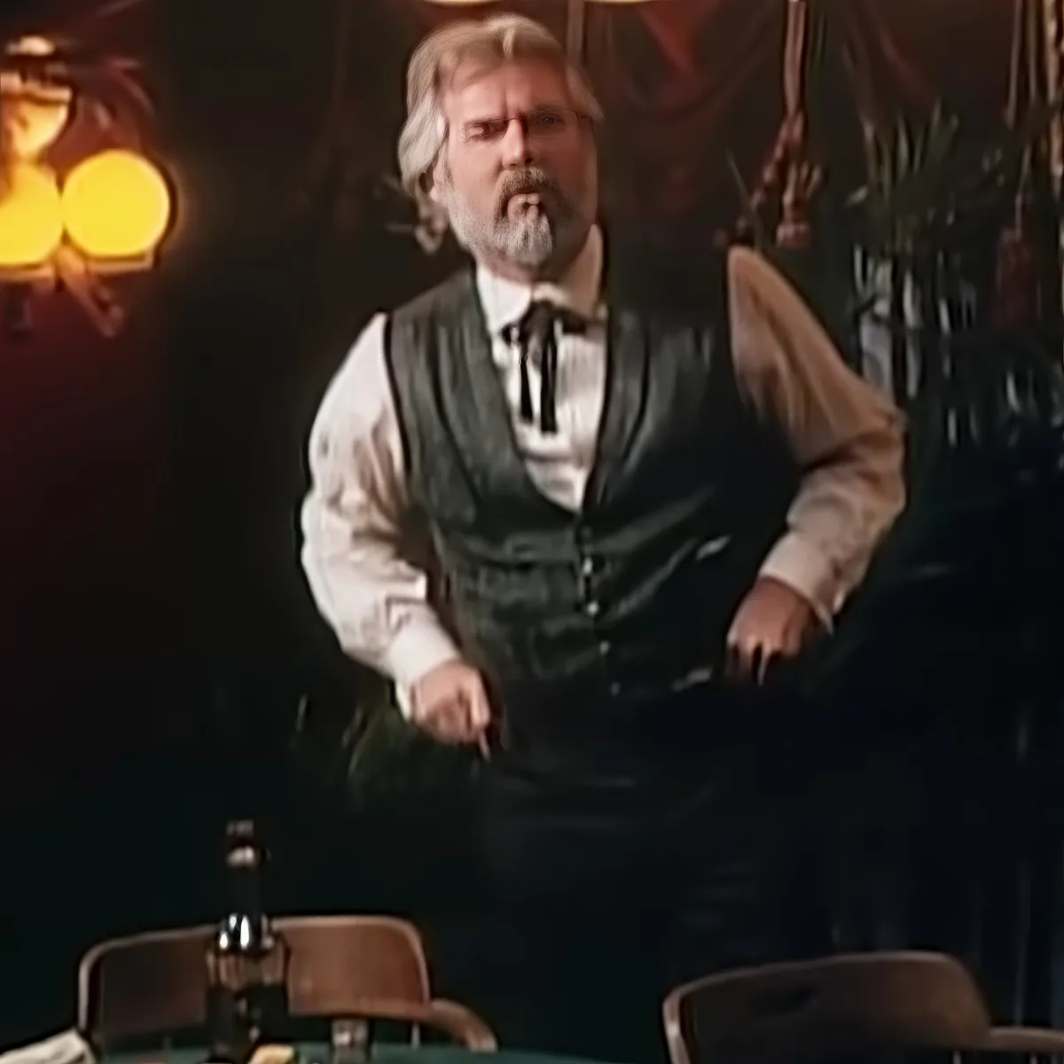 A man in a white shirt, black vest, and bow tie stands in a dimly lit room with chairs and a lamp.