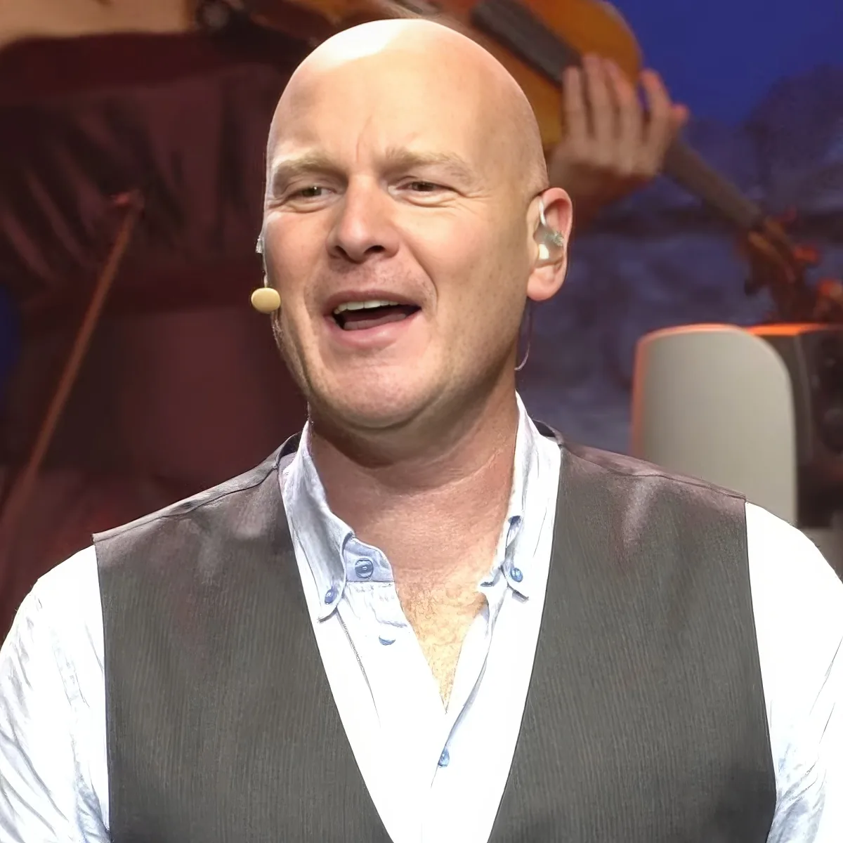 A bald man in a vest and shirt, wearing a microphone, appears to be speaking or singing.