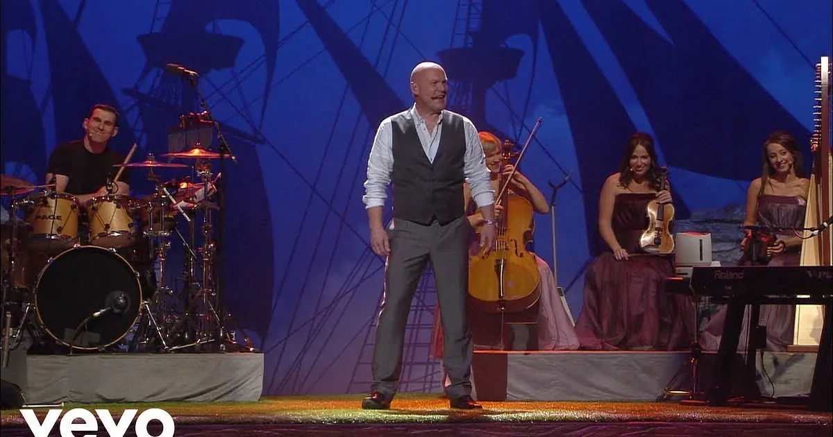 Five men in vests stand on stage, surrounded by musicians and a backdrop of ships and stone walls.