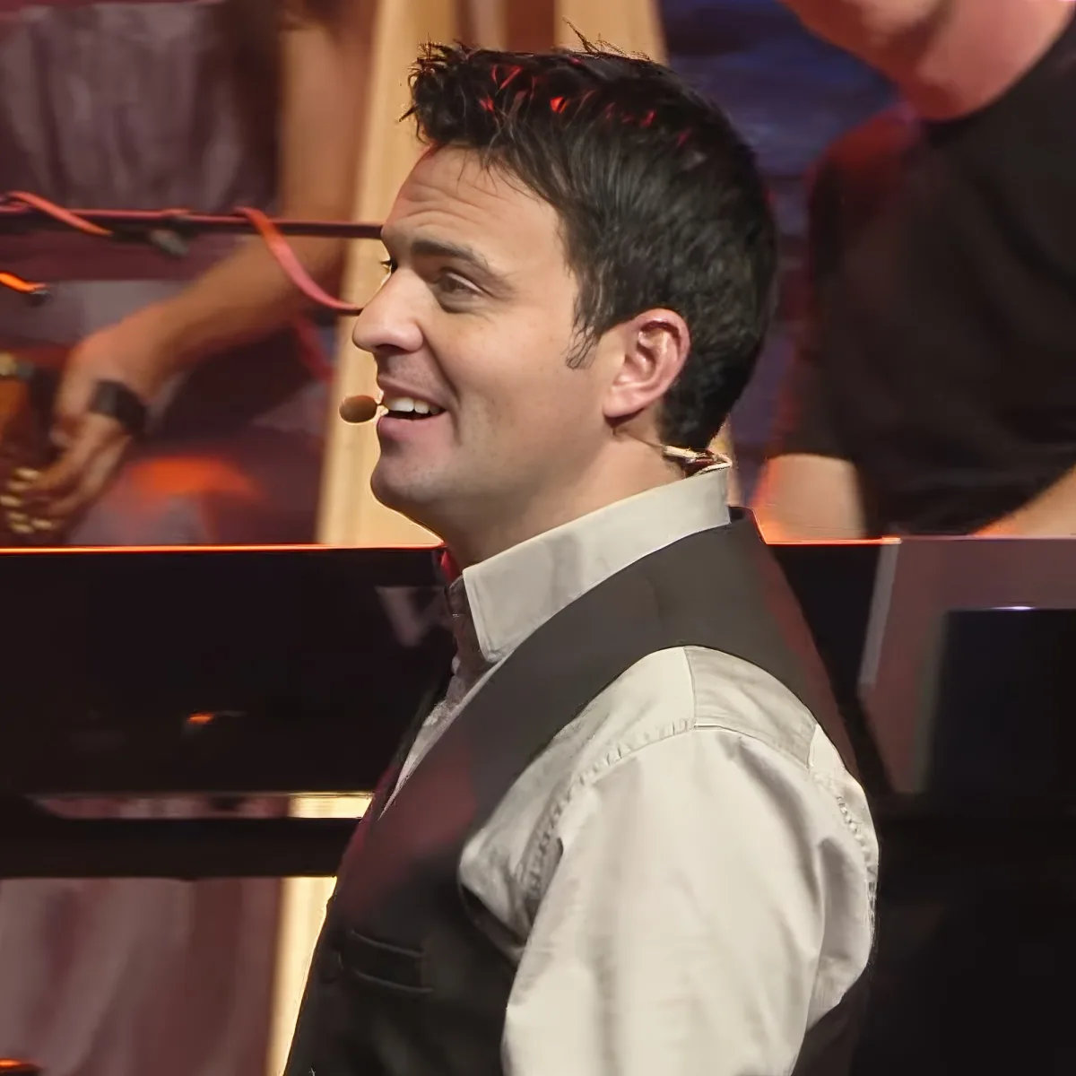 A man in a light shirt and dark vest smiles while wearing a microphone, with a blurred background of a person and musical...