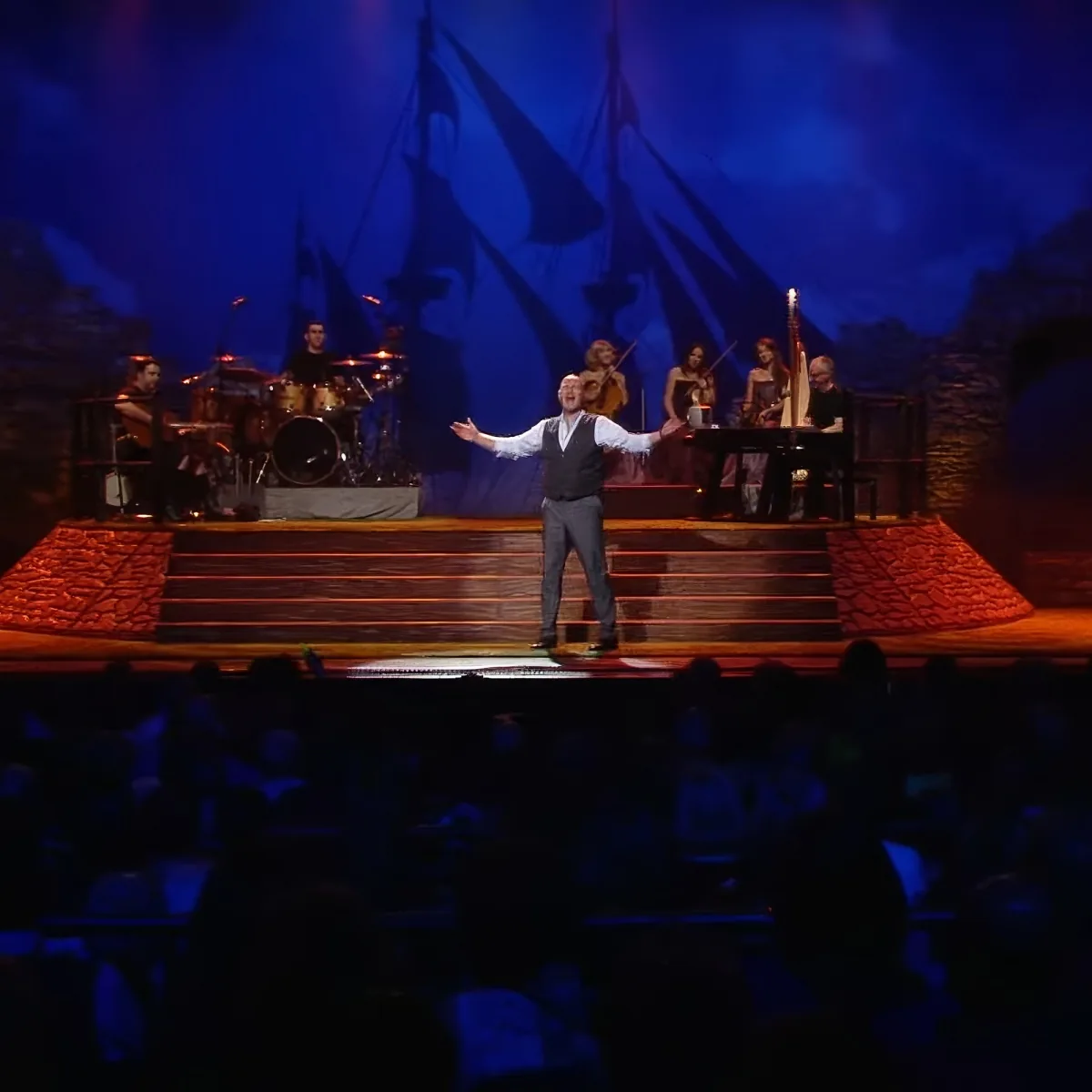A man in a vest stands on stage with arms outstretched, surrounded by musicians and a backdrop of sails.
