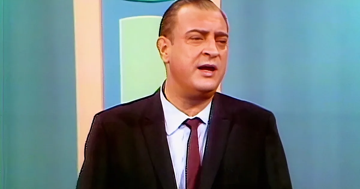 A man in a suit and tie appears to be speaking or reacting, with a background of blue and green hues.