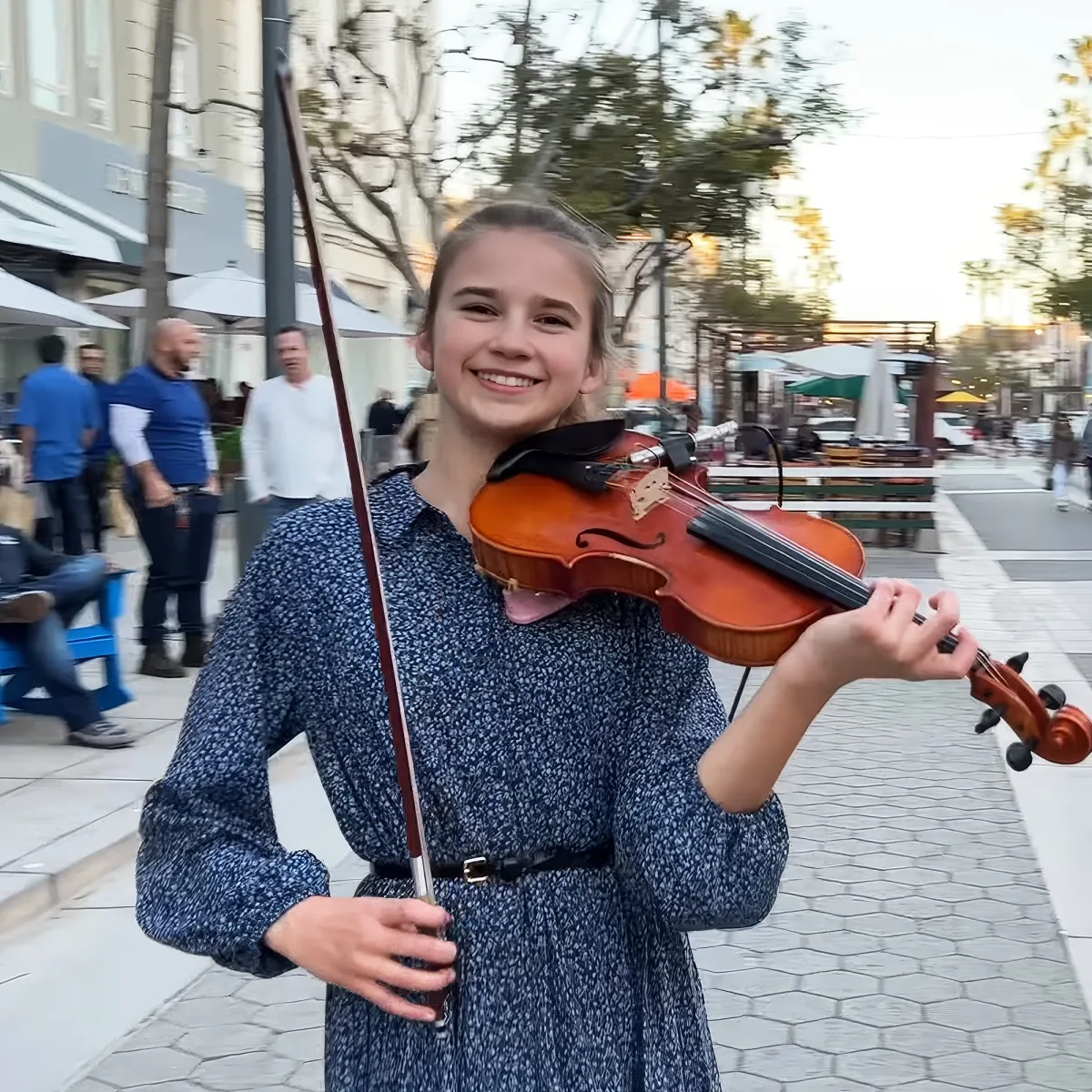 A girl in a blue dress holds a violin, smiling, with a bustling outdoor setting in the background.