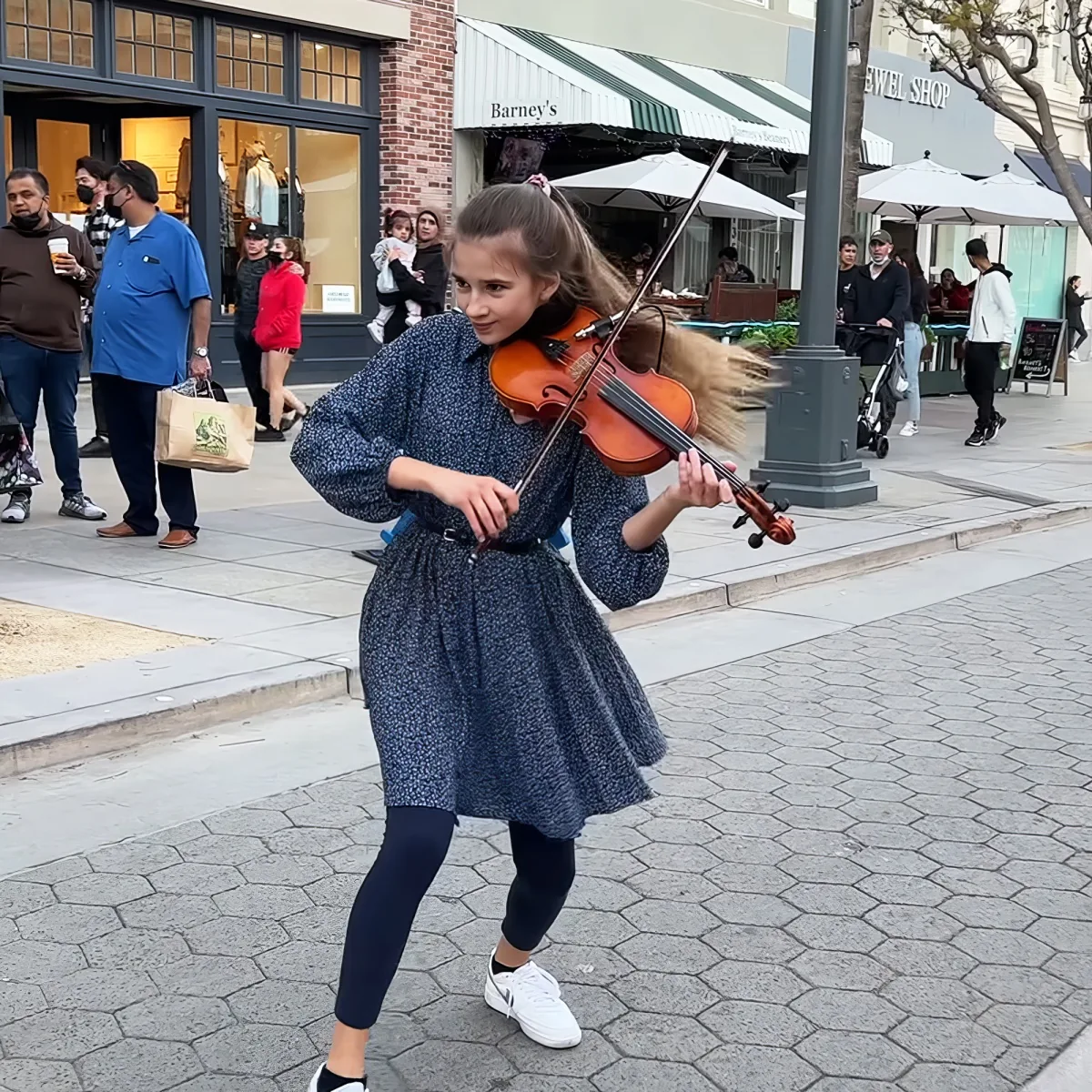 A girl in a blue dress plays the violin on a city sidewalk, surrounded by people and shops.