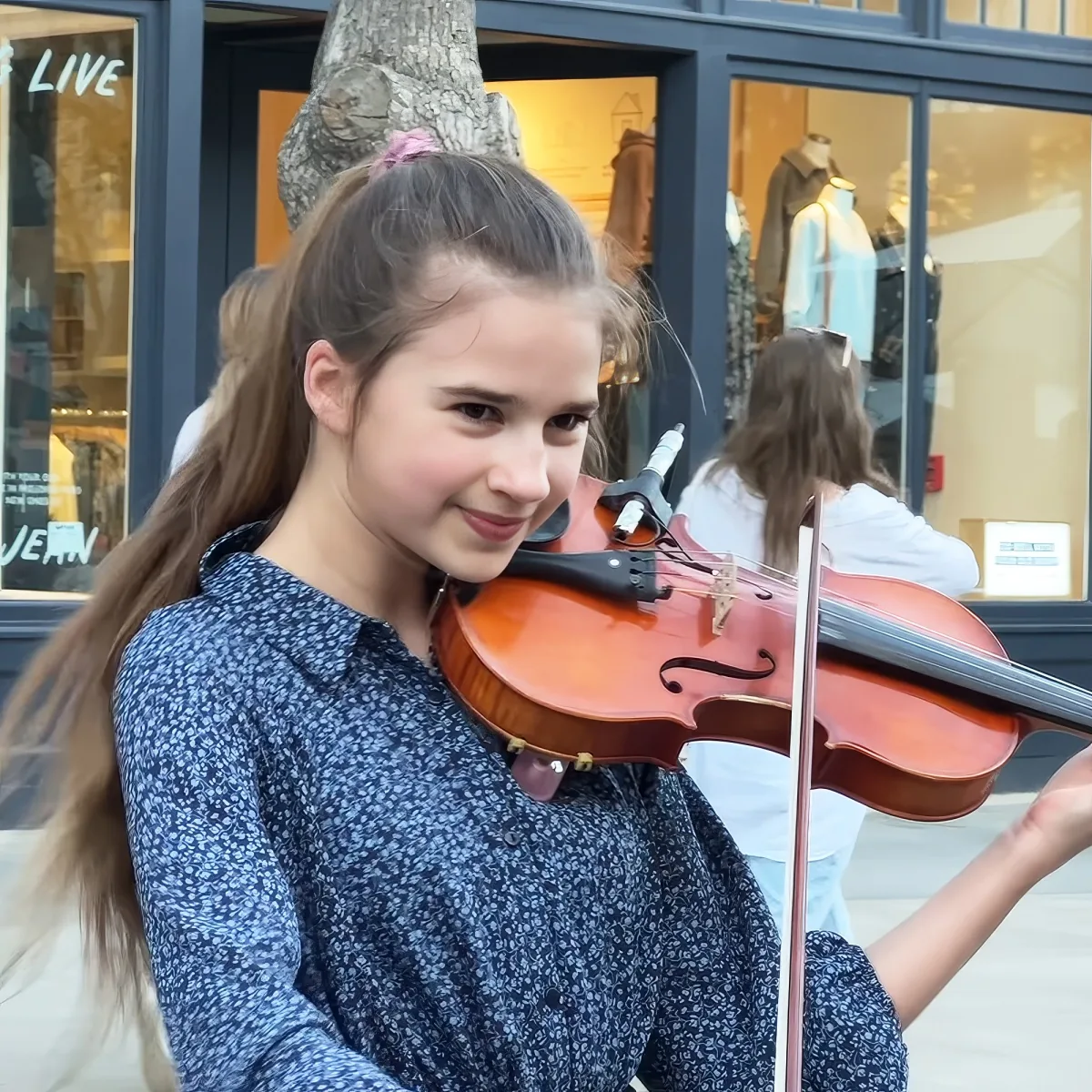 A girl in a blue floral shirt smiles while holding a violin, with a tree and store in the background.