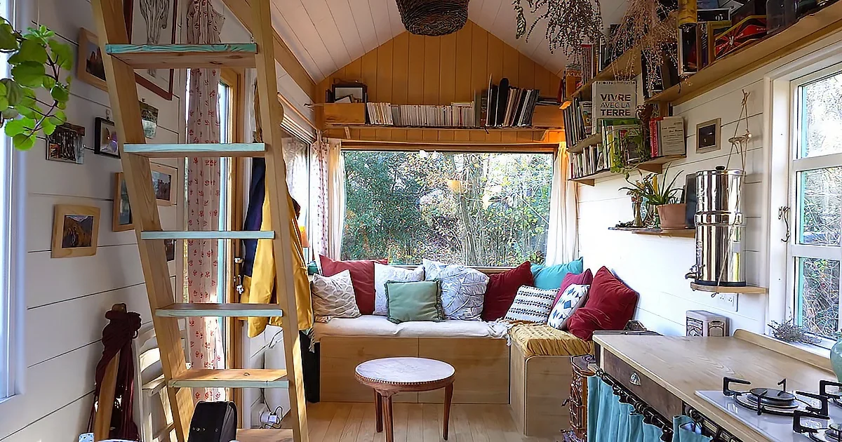 A cozy, well-lit cabin interior with a wooden ladder, colorful cushions, and a view of greenery outside.
