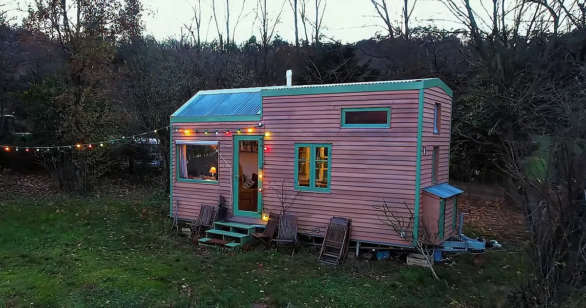 A pink and teal tiny house with string lights and outdoor furniture sits on a grassy area surrounded by trees.
