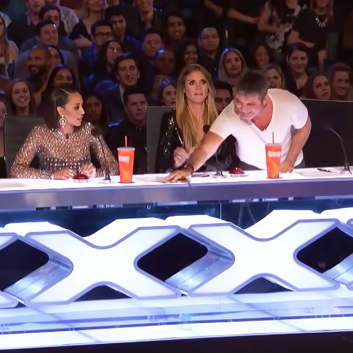 Simon Cowell leans over the table, gesturing while seated with two other judges at a panel.