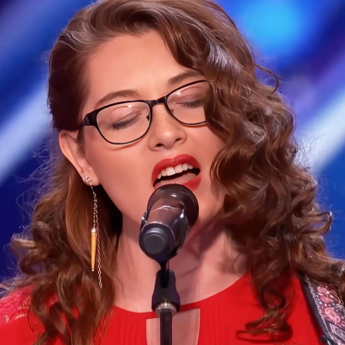 A woman with curly hair, glasses, and red lipstick sings into a microphone, wearing a red top.