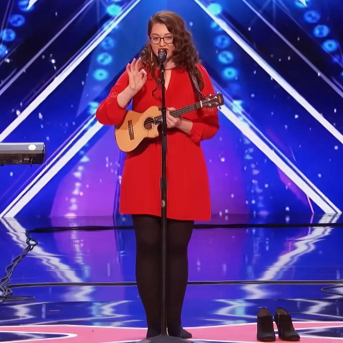 A woman in a red coat and glasses plays a ukulele on stage with a microphone and a blue, illuminated background.