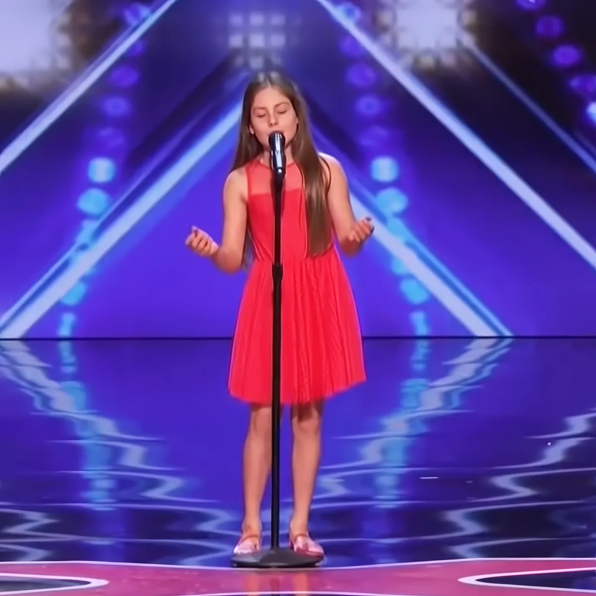 A young girl in a red dress sings on stage with a microphone, hands open, against a purple-lit backdrop.