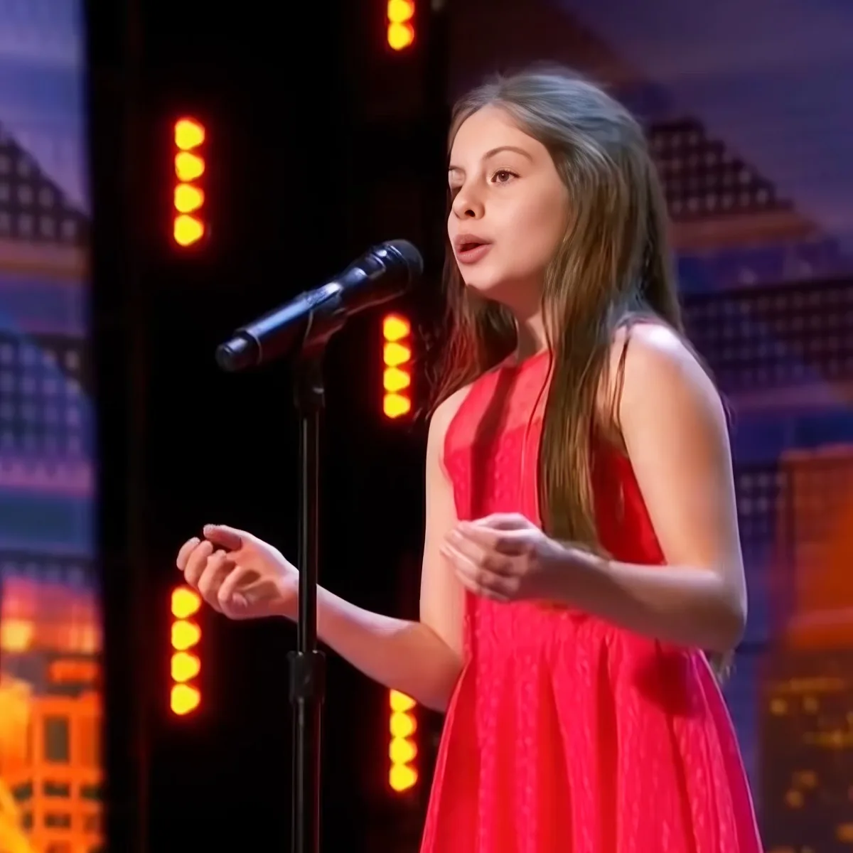 A young girl in a red dress sings into a microphone on a stage with cityscape lights.