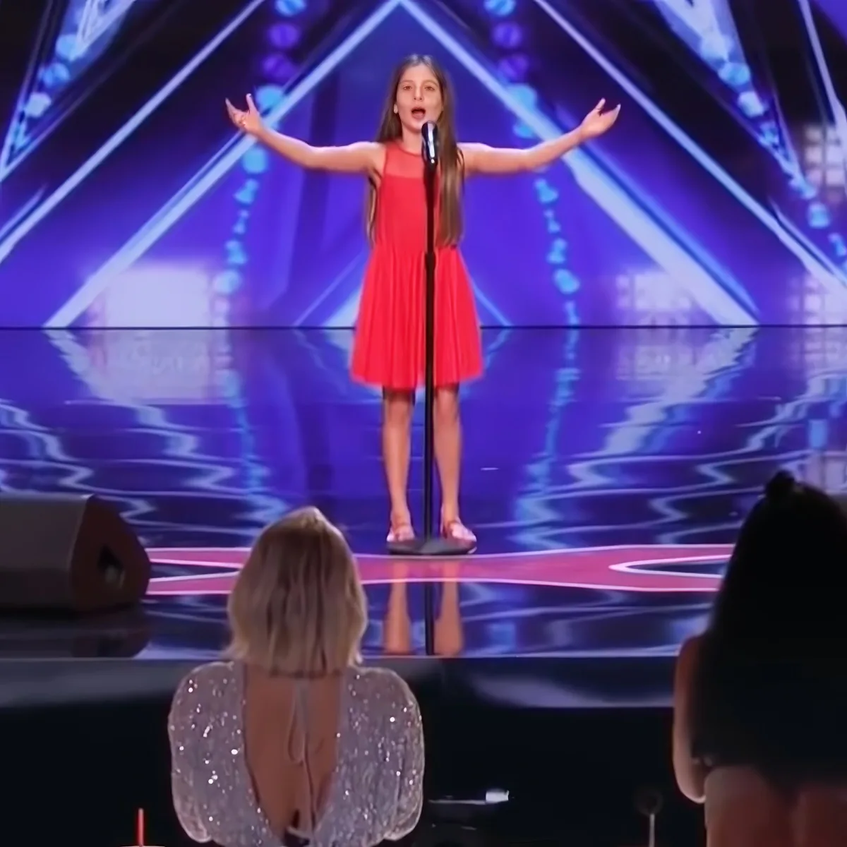 A young girl in a red dress sings passionately on stage with her arms outstretched.