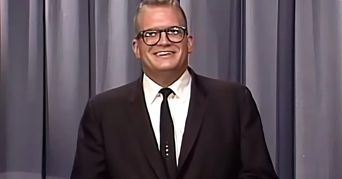 A man in a black suit and white shirt smiles while wearing glasses against a gray curtain backdrop.