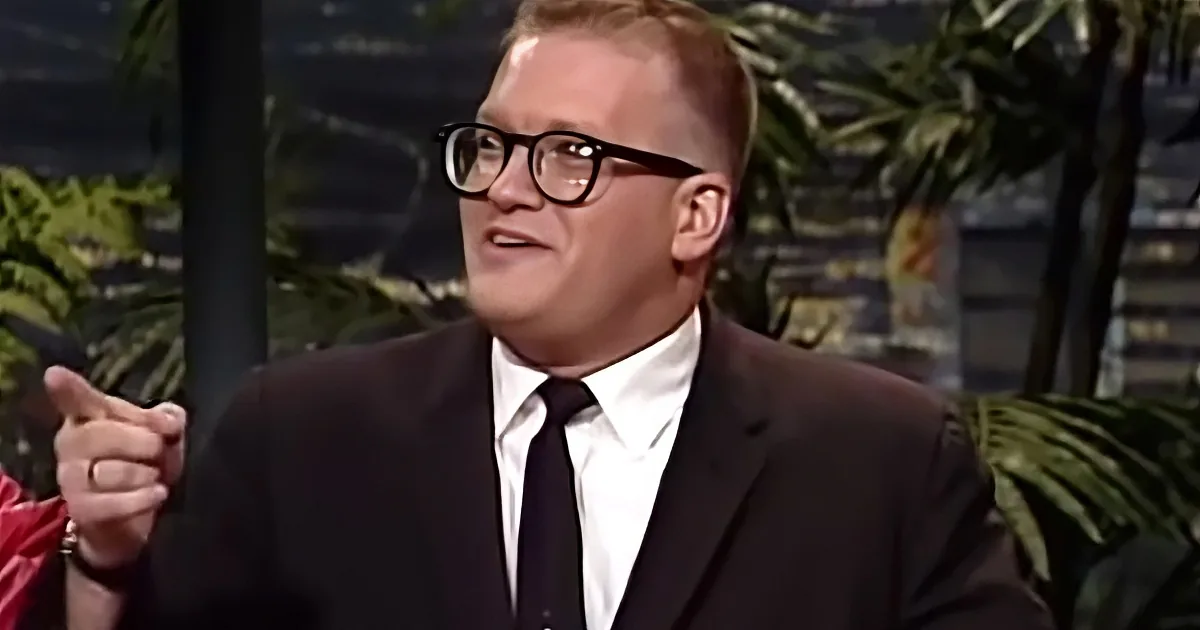 A man in a suit and glasses gestures while speaking, with a tropical backdrop.