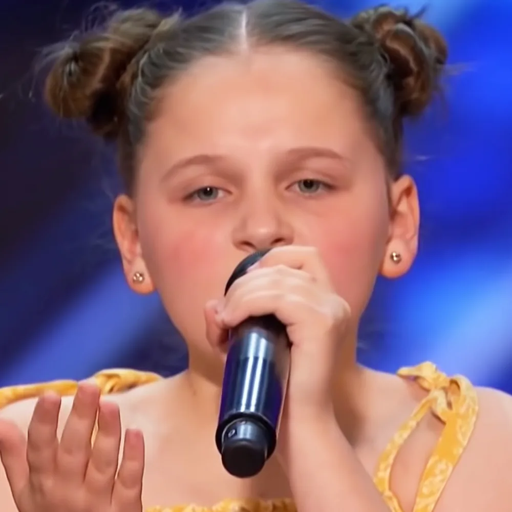 A young girl with pigtails in a yellow top sings into a microphone, her hand raised.