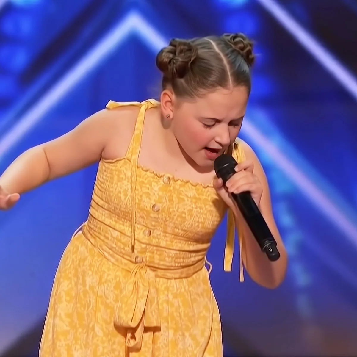 A young girl in a yellow dress sings into a microphone on stage, her hair styled in braids.