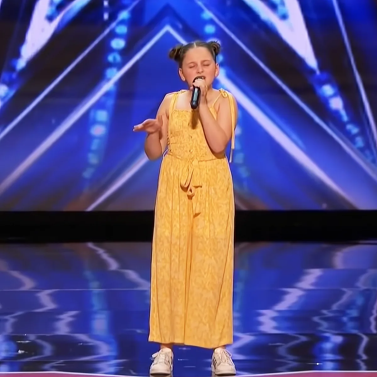 A young girl in a yellow dress sings on stage with a microphone, her hair in pigtails.