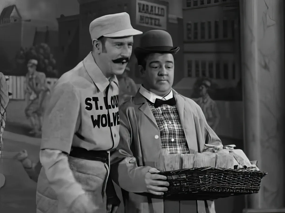 Two men in vintage attire, one holding a basket, standing in front of a hotel backdrop.
