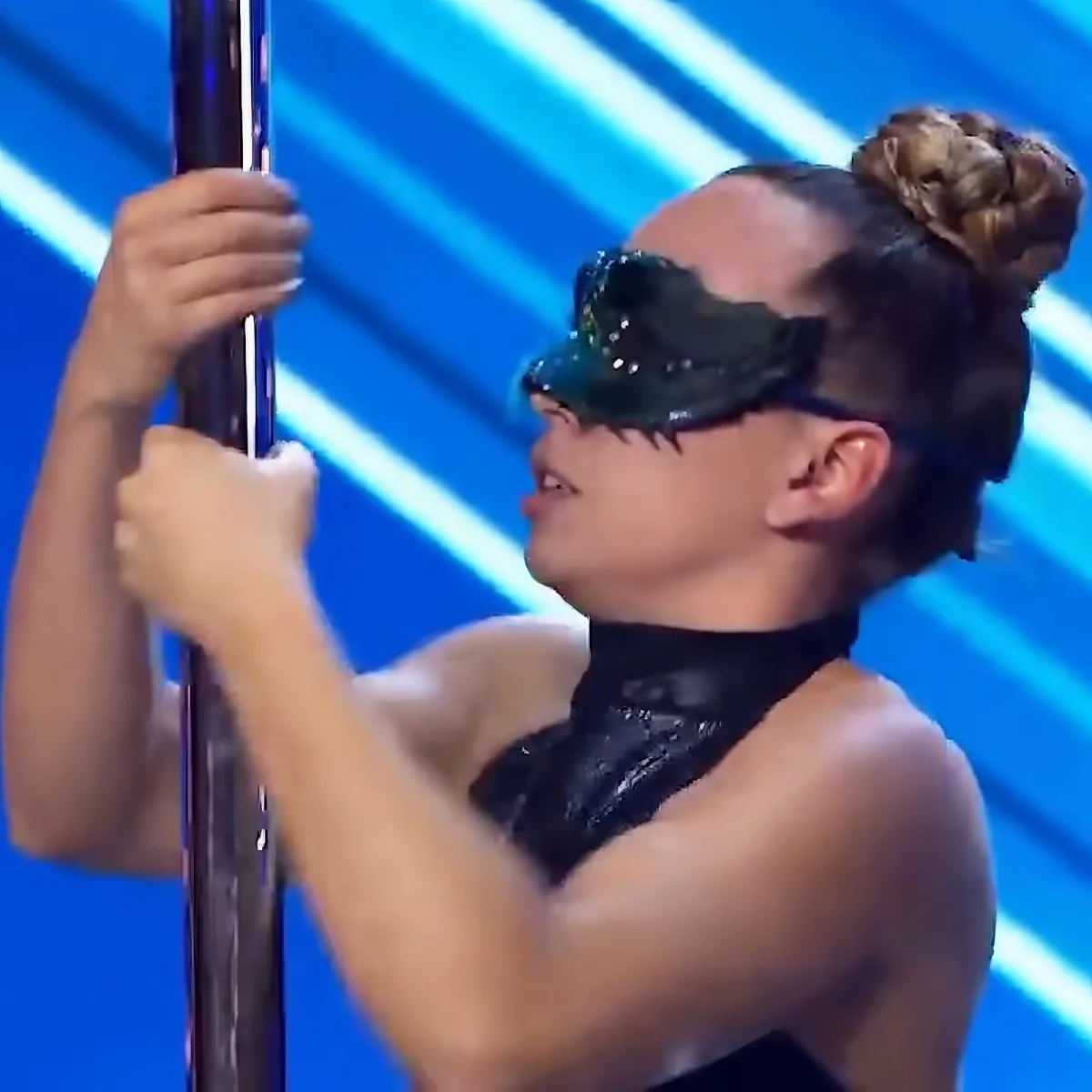 A person in a black outfit and mask is gripping a pole, performing a pole dance move.