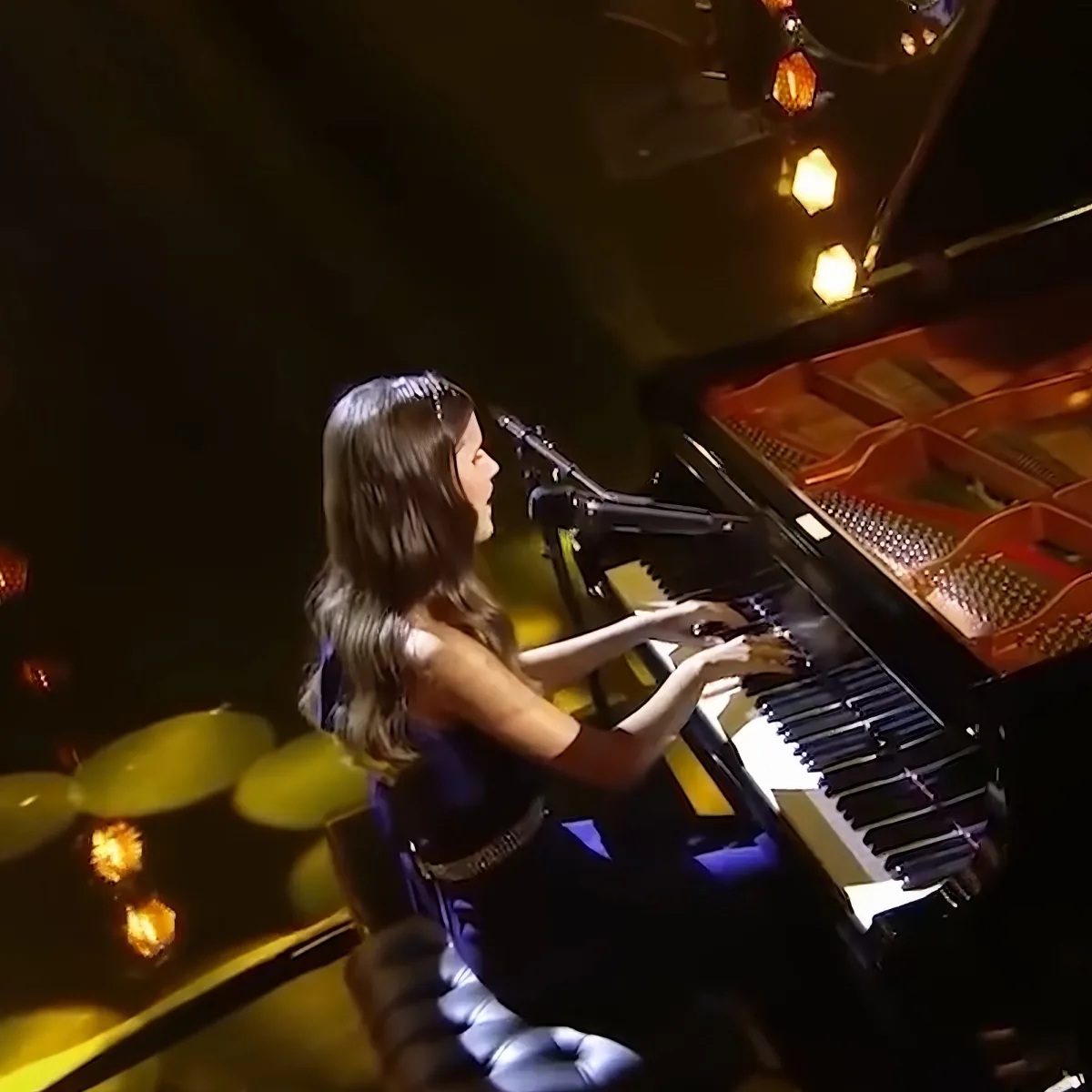 A woman in a dark dress plays a grand piano under warm stage lighting.