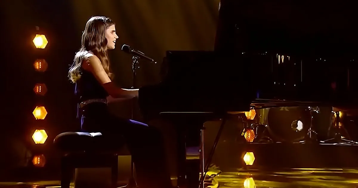A woman sings into a microphone while playing a piano on a dimly lit stage with warm lighting.