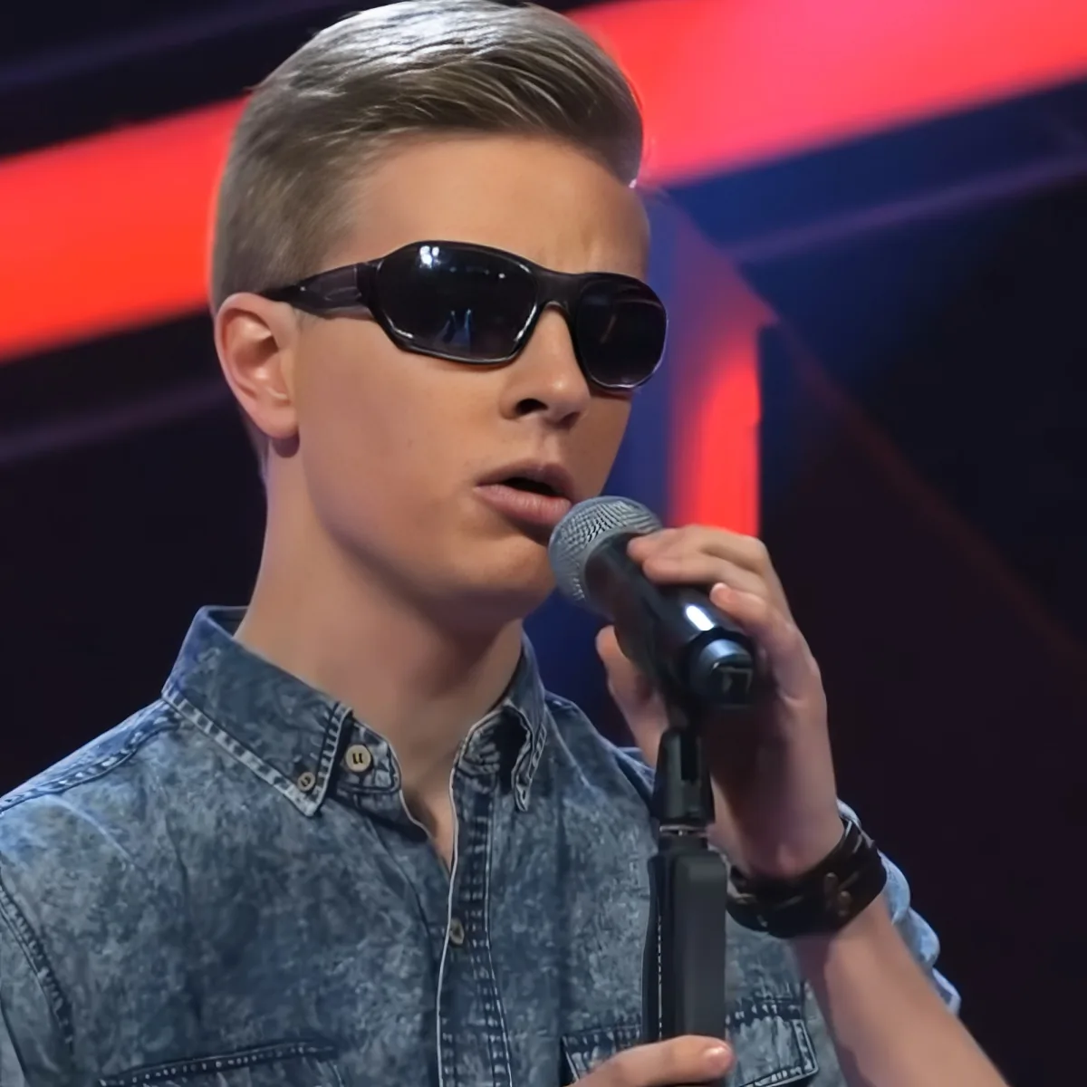 A young man in a denim shirt and sunglasses sings into a microphone on stage.