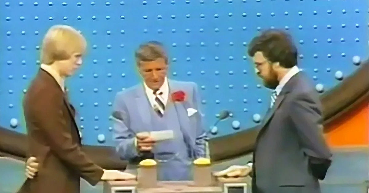 A man in a light blue suit stands behind a desk with microphones, flanked by two other men in suits, in front of a blue...