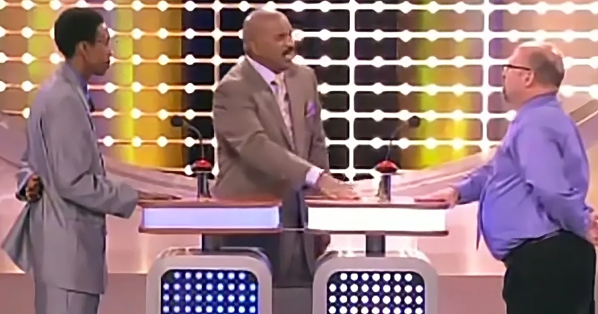 A man in a suit shakes hands with another man at a game show set with colorful lights.