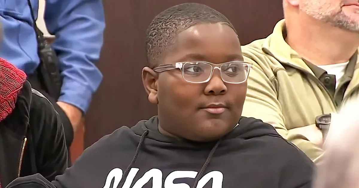 A young boy wearing glasses and a black hoodie with "NASA" on it sits in a courtroom.