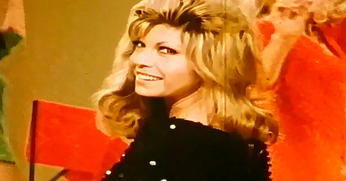 A smiling woman with blonde hair, wearing a black outfit, sits in front of a red background.